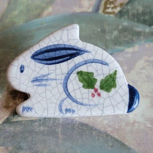 May include: A white ceramic rabbit figurine with blue and green painted details. The rabbit has a cracked glaze and a holly leaf design on its side.