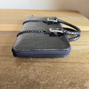 May include: A black rectangular wallet with a crocodile-embossed texture and a zipper closure. The wallet features two braided handles attached with silver-toned metal buckles on the top surface, providing a structured and functional design.