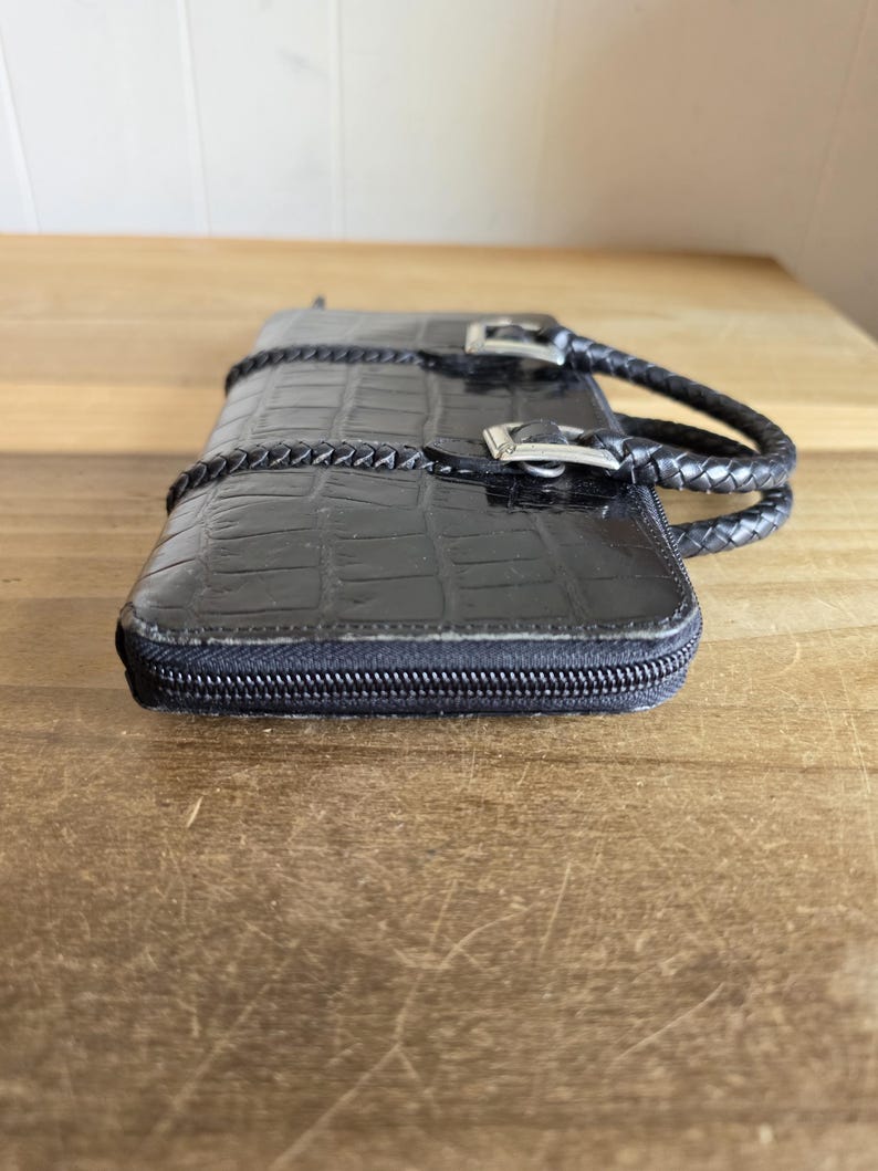 May include: A black rectangular wallet with a crocodile-embossed texture and a zipper closure. The wallet features two braided handles attached with silver-toned metal buckles on the top surface, providing a structured and functional design.