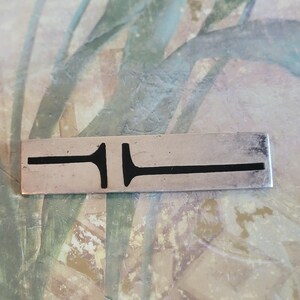 May include: A silver bar pendant with a black abstract design. The pendant is rectangular and has a smooth surface.