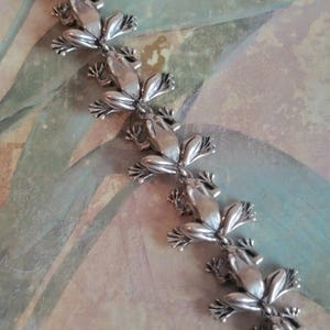 May include: A silver-toned bracelet featuring a series of frog charms. Each frog is detailed with textured skin and outstretched limbs. The bracelet is displayed against a backdrop of muted green and blue hues.
