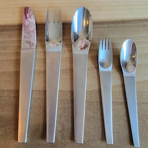 May include: A set of four silver-toned flatware pieces, including a knife, fork, spoon, and a smaller fork. The pieces are arranged on a wooden surface.