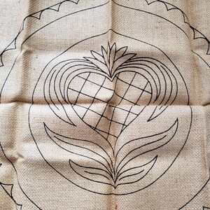 May include: A burlap fabric with a black outline of a heart with a floral design inside. The heart is surrounded by a circle with a scalloped edge.