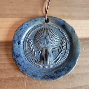 May include: A round, blue ceramic ornament with a wheat sheaf design. The ornament has a textured surface and a leather cord for hanging. The wheat sheaf is surrounded by a laurel wreath.
