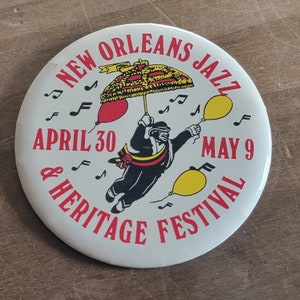 May include: A white button with red and black text that reads "New Orleans Jazz & Heritage Festival" with a black and white illustration of a person holding an umbrella with red, yellow, and white balloons. The text also reads "April 30 - May 9".