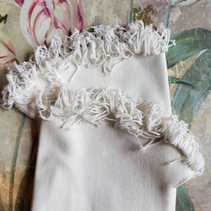 May include: A pair of white fabric gloves with a fringe trim. The gloves are folded and displayed against a floral background.