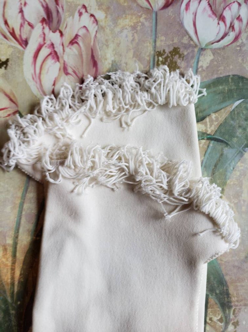 May include: A pair of white fabric gloves with a fringe trim. The gloves are folded and displayed against a floral background.