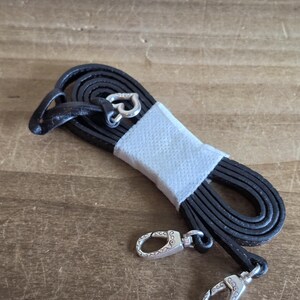 May include: A coiled black leather strap featuring silver-toned metal lobster claw clasps at each end. The strap is secured in the center with a white paper band and includes a small silver-toned heart-shaped charm attached near one of the clasps.