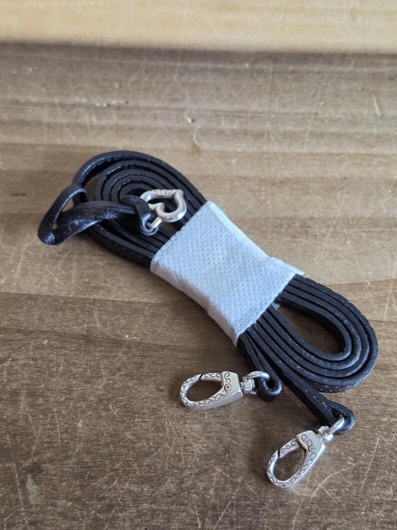 May include: A coiled black leather strap featuring silver-toned metal lobster claw clasps at each end. The strap is secured in the center with a white paper band and includes a small silver-toned heart-shaped charm attached near one of the clasps.