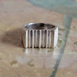 May include: A silver ring with a rectangular band. The ring features vertical, ridged detailing around the entire band. The ring is shown on a marble surface.