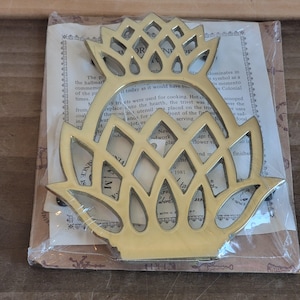 May include: A gold-toned pineapple-shaped trivet, a traditional kitchen utensil used to protect surfaces from hot cookware. The trivet is packaged in a clear plastic bag with a printed card that describes its history and use.