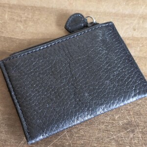 May include: A rectangular black leather coin purse with a textured, pebbled surface. The accessory features a top zipper closure with a small, matching black leather pull tab attached to a silver-toned metal ring, providing a secure opening.