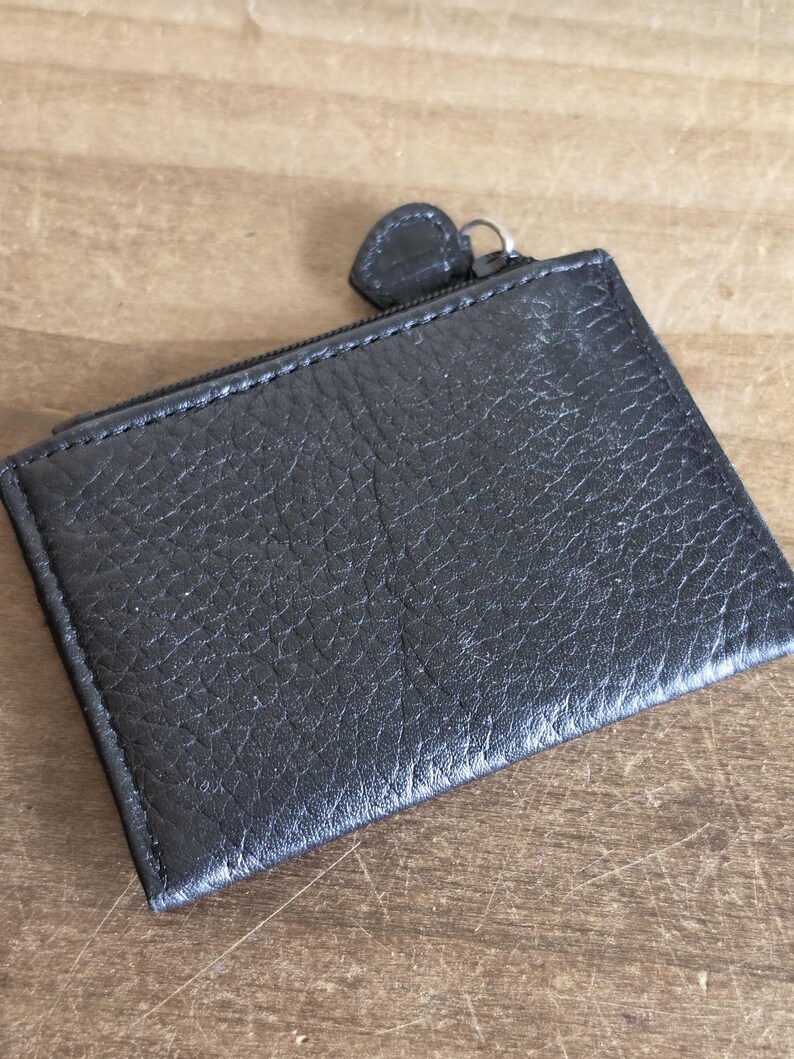 May include: A rectangular black leather coin purse with a textured, pebbled surface. The accessory features a top zipper closure with a small, matching black leather pull tab attached to a silver-toned metal ring, providing a secure opening.