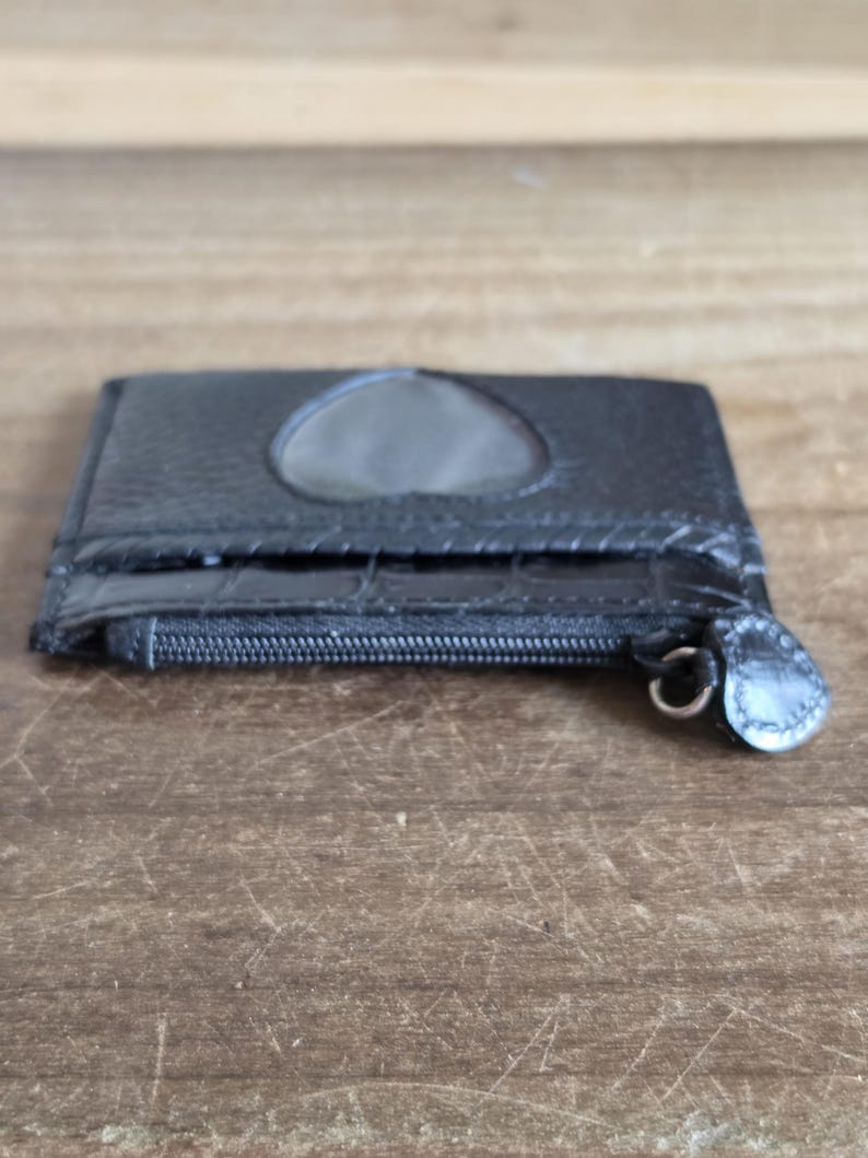 May include: A black card holder with a textured surface, featuring a heart-shaped cutout window on the front and a zippered compartment. The zipper pull is shaped like a small heart, and the front pocket has a glossy, embossed crocodile-like pattern.