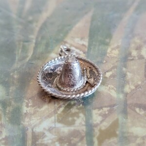 May include: A silver charm in the shape of a sombrero. The sombrero has a wide brim and a tall crown. The charm is detailed with a rope trim around the brim.