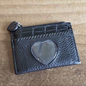 May include: A black leather card holder featuring a textured finish, a top zipper closure with a heart-shaped pull, and a central heart-shaped clear plastic window. The upper section has a crocodile-embossed pattern with a diagonal stitched detail.