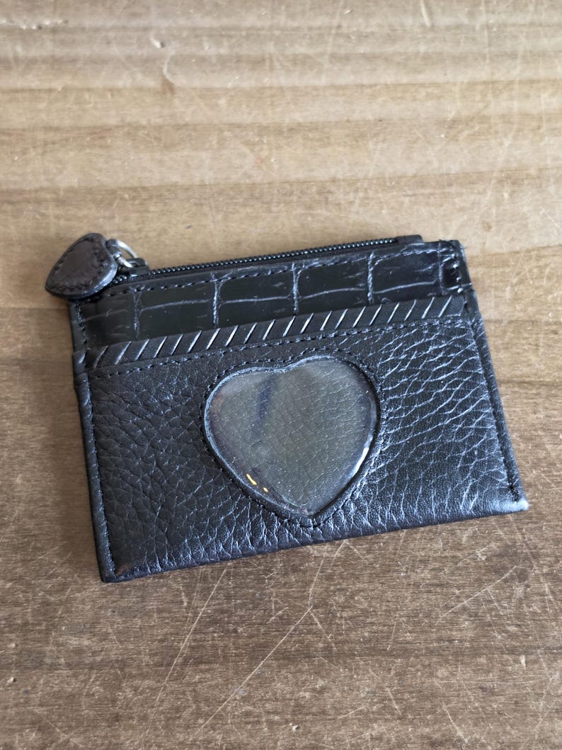 May include: A black leather card holder featuring a textured finish, a top zipper closure with a heart-shaped pull, and a central heart-shaped clear plastic window. The upper section has a crocodile-embossed pattern with a diagonal stitched detail.