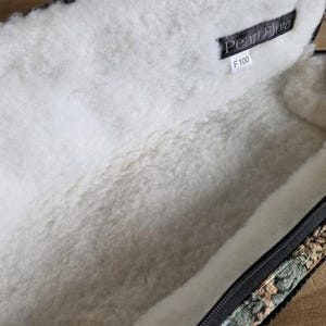 Puede incluir: Estuche de flauta abierto con un interior blanco y suave. El estuche tiene una cremallera negra y un exterior estampado con tonos verdes, marrones y beiges. Una etiqueta negra en el interior dice "Pearl Flute" y "F 100".