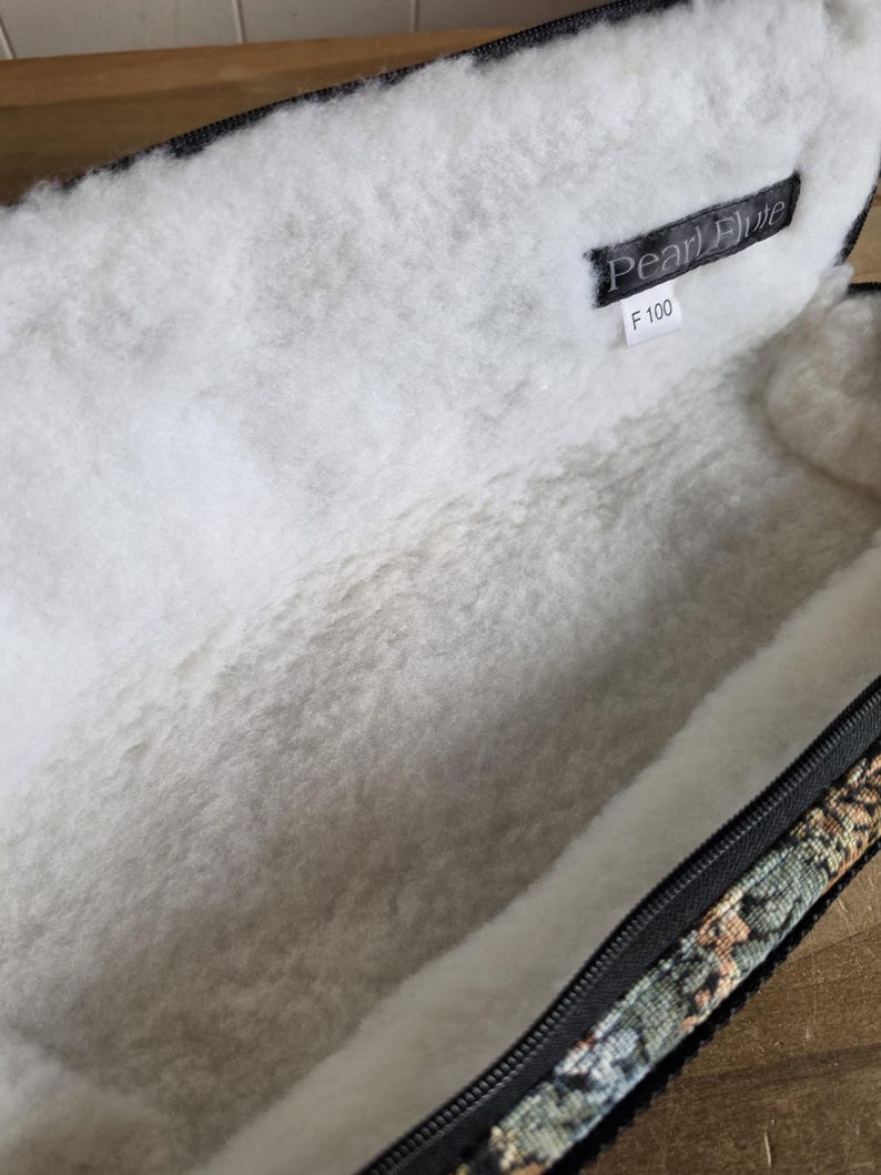 Puede incluir: Estuche de flauta abierto con un interior blanco y suave. El estuche tiene una cremallera negra y un exterior estampado con tonos verdes, marrones y beiges. Una etiqueta negra en el interior dice "Pearl Flute" y "F 100".