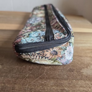 Puede incluir: Un estuche rectangular con estampado floral y cierre de cremallera negro. El estuche presenta un dise&ntilde;o tipo tapiz con una mezcla de colores, incluyendo rosa, azul y verde. El estuche est&aacute; sobre una superficie de madera.