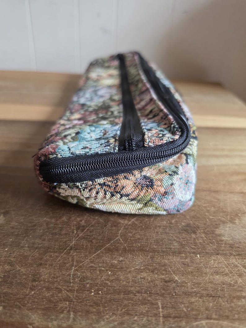 Puede incluir: Un estuche rectangular con estampado floral y cierre de cremallera negro. El estuche presenta un dise&ntilde;o tipo tapiz con una mezcla de colores, incluyendo rosa, azul y verde. El estuche est&aacute; sobre una superficie de madera.