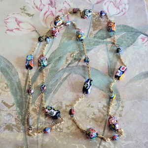 May include: A long gold chain necklace with colorful, patterned glass beads. The beads are a mix of oval and rectangular shapes and feature a variety of floral and geometric designs.