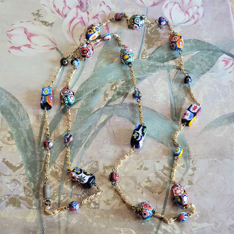 May include: A long gold chain necklace with colorful, patterned glass beads. The beads are a mix of oval and rectangular shapes and feature a variety of floral and geometric designs.