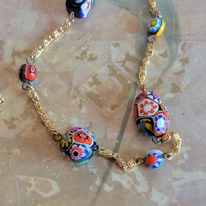 May include: A gold chain necklace with colorful, patterned glass beads. The beads are oval and round in shape and feature a variety of colors, including red, blue, yellow, and green.