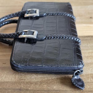 May include: A black rectangular wallet with a glossy crocodile-embossed texture. It features two braided black handles attached with silver-toned metal buckles. The wallet is secured with a black zipper closure that includes a matching black pull tab.