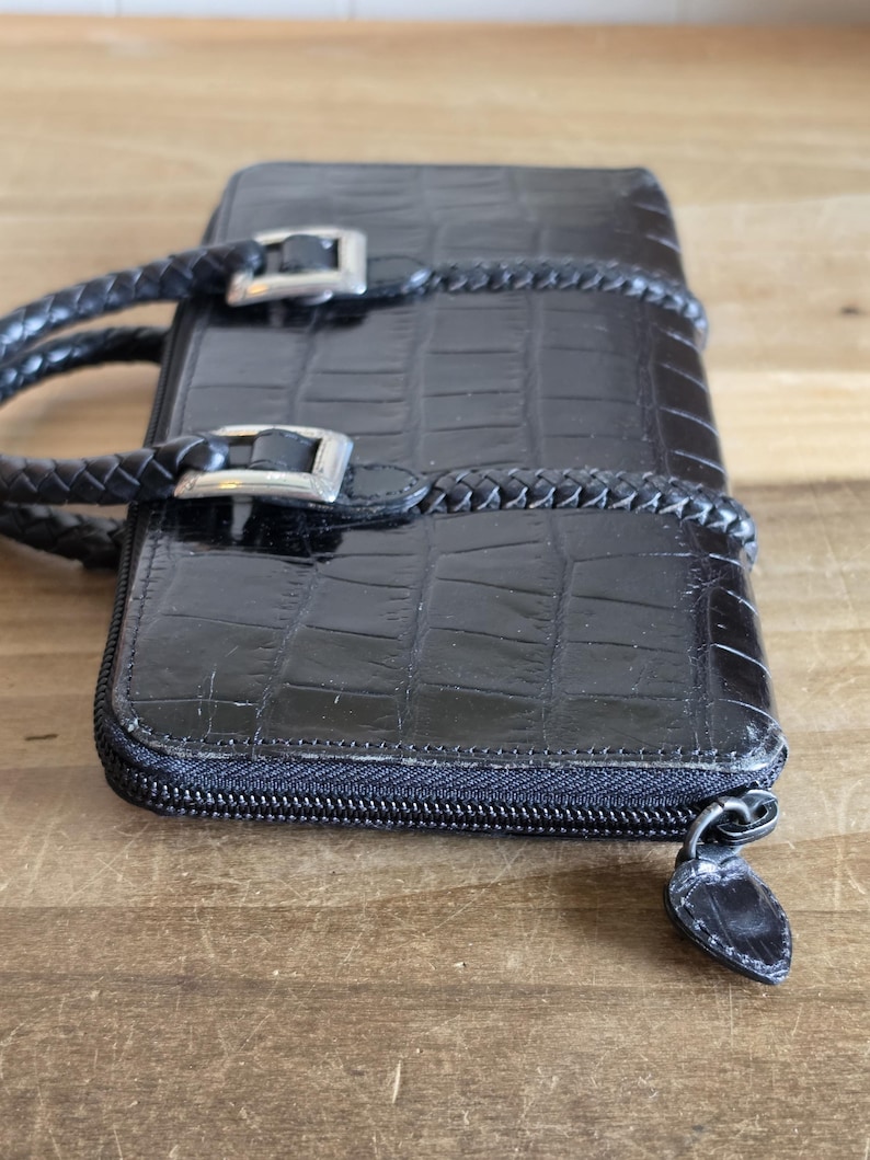 May include: A black rectangular wallet with a glossy crocodile-embossed texture. It features two braided black handles attached with silver-toned metal buckles. The wallet is secured with a black zipper closure that includes a matching black pull tab.