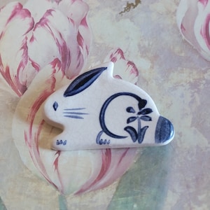 May include: A white ceramic rabbit figurine with blue and black painted details. The rabbit is sitting on a floral background.