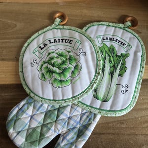 May include: A set of kitchen textiles including a round pot holder with a green lettuce illustration and the text LA LAITUE, an oval pot holder with a green vegetable illustration and the text LABLETTE, and a quilted oven mitt with a green and blue plaid pattern.