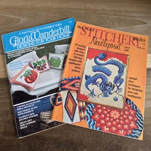 May include: Two vintage craft magazines titled Gloria Vanderbilt Designs for Your Home and McCall's Stitchery Series Needlepoint IV. The covers feature colorful needlepoint projects including a blue dragon, floral patterns, and geometric designs.