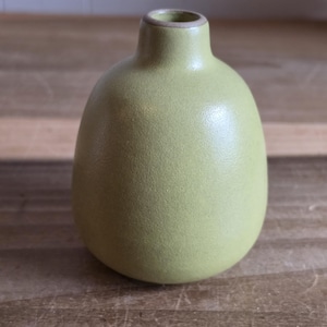 May include: A small, light green ceramic vase with a rounded body and a narrow neck. The vase has a textured surface and sits on a wooden surface. The vase is a decorative item.