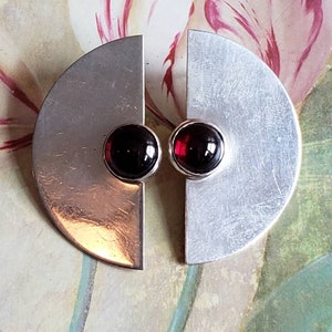 May include: A pair of silver earrings with a geometric design. Each earring features a half-circle shape with a red gemstone set in the center.