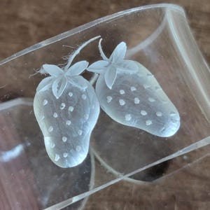 May include: Close-up of a clear glass object with two etched strawberries. The strawberries have detailed leaves and seeds, and are positioned on the side of the glass. The glass is partially visible, showing its curved shape.