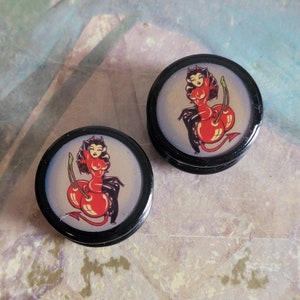 May include: Pair of black acrylic ear gauges with a red and black illustration of a woman with devil horns sitting on two cherries.