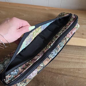 Puede incluir: Estuche de l&aacute;pices abierto de tapiz floral con cremallera negra. El interior es negro. El estuche est&aacute; abierto, revelando el forro interior. El estampado floral incluye colores rosa, verde y beige.