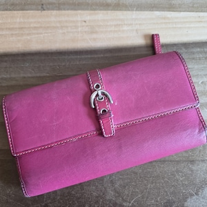 May include: A magenta leather wallet with a silver-tone buckle closure. The wallet features white stitching and a small tab on the side. The rectangular wallet is closed and appears to be made of a smooth leather material.