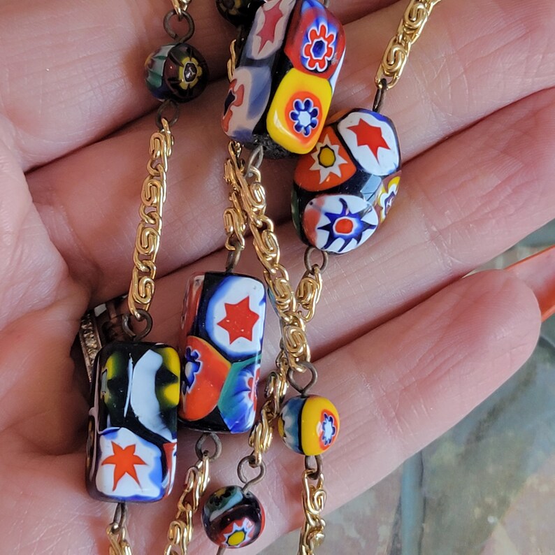 May include: A gold chain necklace with colorful glass beads. The beads are round and rectangular, and they feature a variety of patterns, including stars, flowers, and geometric shapes. The beads are in shades of red, yellow, blue, and green.