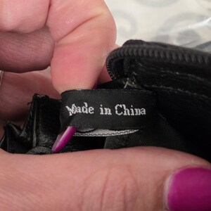 May include: A black fabric tag with white text that reads Made in China, attached to the interior of a black item with a zipper. The tag is rectangular and positioned horizontally, showing clear white lettering against the dark background.