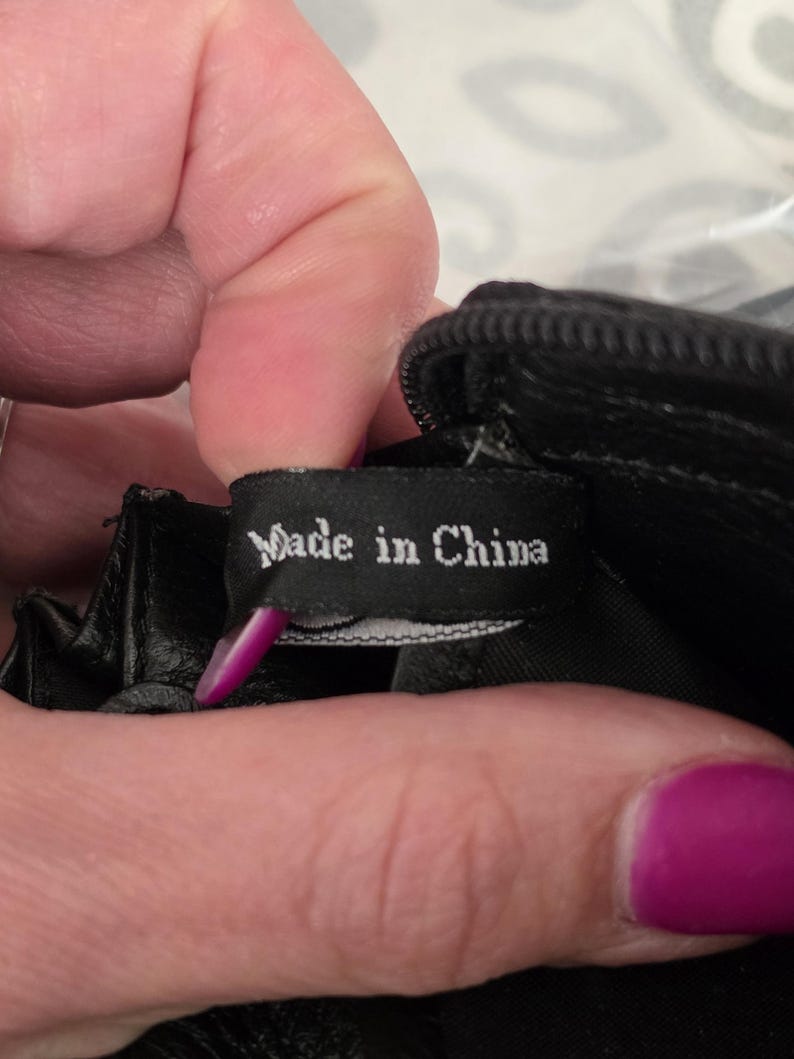 May include: A black fabric tag with white text that reads Made in China, attached to the interior of a black item with a zipper. The tag is rectangular and positioned horizontally, showing clear white lettering against the dark background.