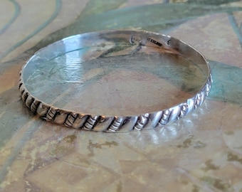 May include: A silver-colored bangle bracelet with a textured design. The bracelet has a repeating pattern of diagonal lines and small rectangular shapes. The metal has a polished finish, reflecting light. The bracelet is a piece of jewelry.
