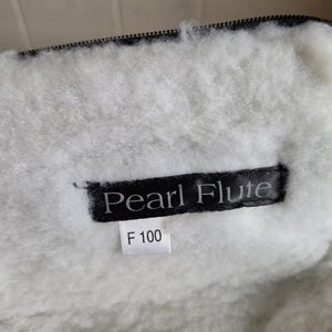 Puede incluir: Primer plano de un forro interior blanco y esponjoso con una cremallera negra. Se ve una etiqueta negra con el texto "Pearl Flute", junto con una etiqueta m&aacute;s peque&ntilde;a que dice "F 100". El forro parece estar hecho de un material suave y afelpado.