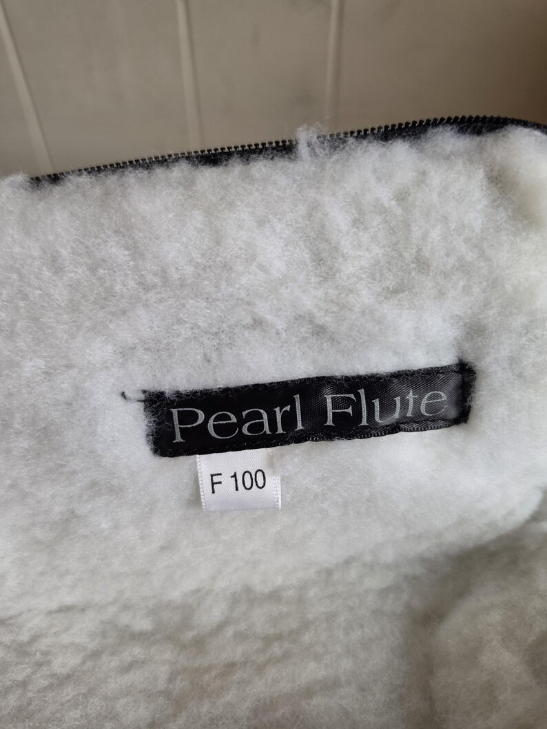 Puede incluir: Primer plano de un forro interior blanco y esponjoso con una cremallera negra. Se ve una etiqueta negra con el texto "Pearl Flute", junto con una etiqueta m&aacute;s peque&ntilde;a que dice "F 100". El forro parece estar hecho de un material suave y afelpado.