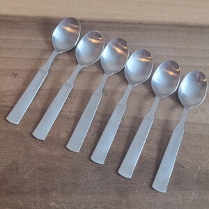 May include: Six stainless steel spoons arranged on a wooden surface. Each spoon has a rounded bowl and a rectangular handle with a decorative pattern near the top. The spoons are evenly spaced and angled.