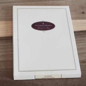 May include: A white box of Victoria's Secret stockings. The box has a cream-colored border and a maroon oval with the text "Victoria's Secret Lingerie for Legs." The bottom of the box reads "STOCKINGS Midnight Black / Medium."