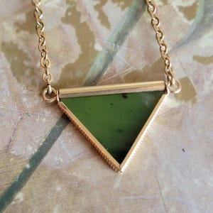 May include: A gold-colored necklace featuring a triangular, dark green stone pendant. The pendant is framed by a gold-colored metal with a textured edge. The necklace chain is also gold-colored and delicate.
