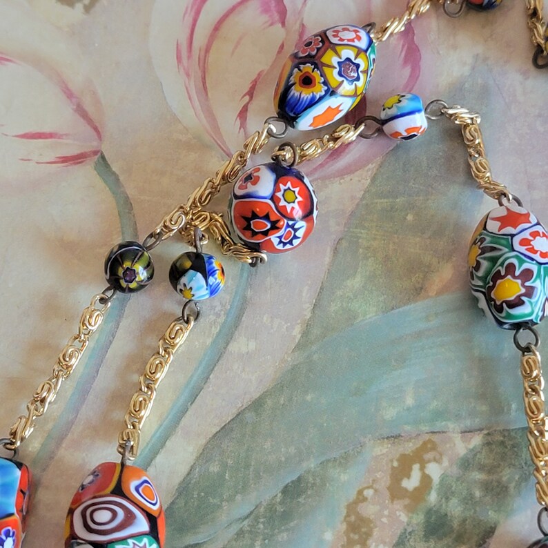 May include: A gold chain necklace with colorful, patterned glass beads. The beads are round and oval shaped and feature floral designs in shades of red, blue, green, yellow, and white.
