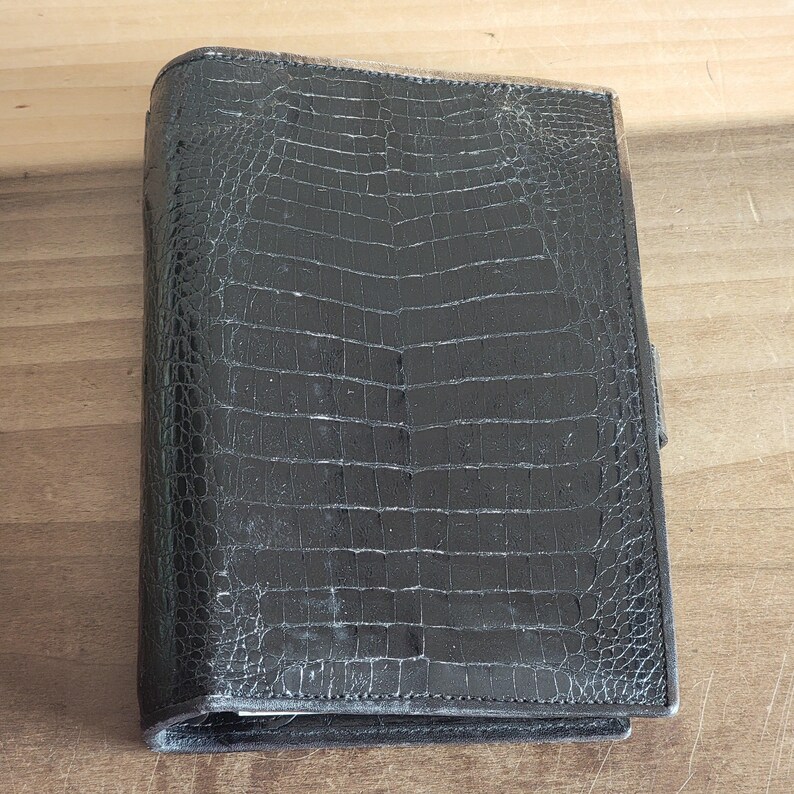 Puede incluir: Un diario encuadernado en cuero negro con un patr&oacute;n de cocodrilo en relieve.